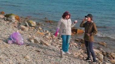 İki kadın, kirliliği önlemek için kumsaldaki atıkları temizliyor. Kadınlar bir fren yapar ve çay içerler. İnsanlar Dünya Günü 'nde okyanusu temizleme girişimine katılıyorlar. Tam boy çekim.