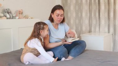 Küçük beyaz kız, aydınlık bir odada yatağında otururken annesiyle okumayı öğreniyor. Evde erken eğitim ve anaokulu gelişimi. Geniş açı.