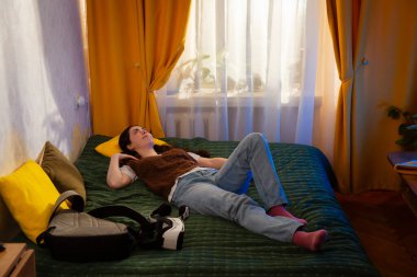 Kadın, yumuşak ışıklı bir odada yüzünde VR bardakları ile yatağında oturuyor ve rahatsızlık gösteriyor. Dijital etkileşime ve yeni teknolojik keşiflere gerçekçi bir tepki. Geniş açı.