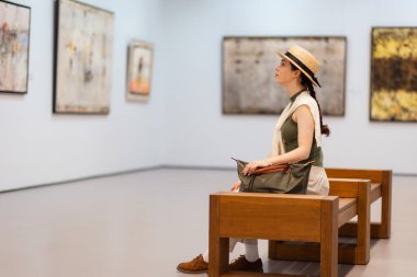 Hasır şapkalı genç bir kadının modern galerideki tabloların sergisine hayran olup bankta oturmasını yan görüyorum. Çağdaş sanat ve kültür eğitimi kavramı.