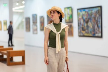 Hasır şapkalı ve zarif elbiseli güzel beyaz bir kadının çağdaş bir sanat galerisini ziyaret edişinin orta fotoğrafı. Müze sergisi ve kültür eğitimi.