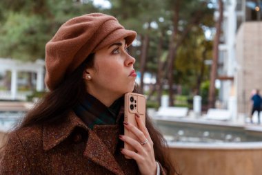 Bej ceketli ve bereli güzel beyaz kadın portresi akıllı telefonunu kullanarak sonbahar şehrinde yürüyor ve rahatlatıcı bir hafta sonu atmosferinin tadını çıkarıyor.