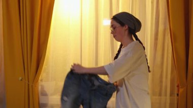 Genç bayan rahat bir yatak odasında oturur ve yatağın üzerinde kıyafet eşyalarını katlar ve düzenler. Yolculuğa hazırlanır. Yan görünüm.