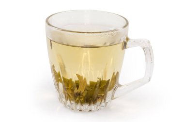 Şeffaf cam bardakta taze demlenmiş yeşil çay fincanın altında beyaz arka planda çay yaprakları var.