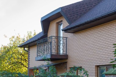 Modern, tipik, sarı tuğlalı bir ev, sahte çelik korkuluklu bir balkon ve gökyüzüne karşı çatının bir parçası.