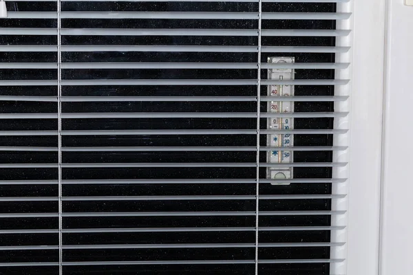 Part of the modern window with window pane covered with water drops and attached outdoor thermometer, inside view across the blinds illuminated from within at night