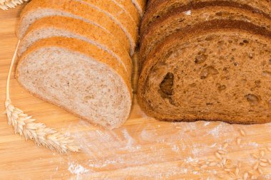 Sliced two oblong loaves of the dark brown bread made from rye and wheat flour and bread with bran among the ears and grain on the cutting board strewn with flour, close-up in selective focus