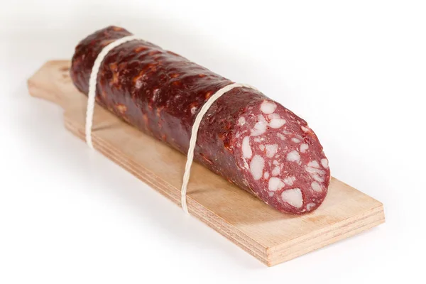 Piece of the smoked-cured sausage made with adding of spinal fat tied to the specially designed narrow wooden cutting board for charcuterie, close-up in selective focus on a white surface