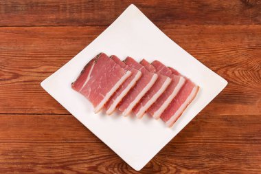 Slices of the dry-cured pork belly on a skin on the white dish square shape on the rustic table, top view
