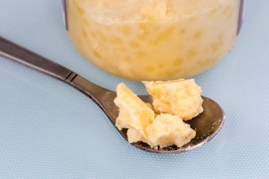 Çay kaşığındaki parçalanmış kakao yağı parçaları mavi bir yüzeyde, aynı kakao yağının küçük cam kavanozuna karşı.