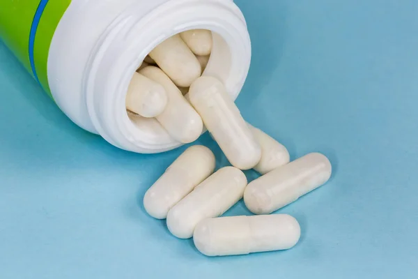 White capsules of the medication partly poured out from container in the form small plastic bottle on the blue surface close-up