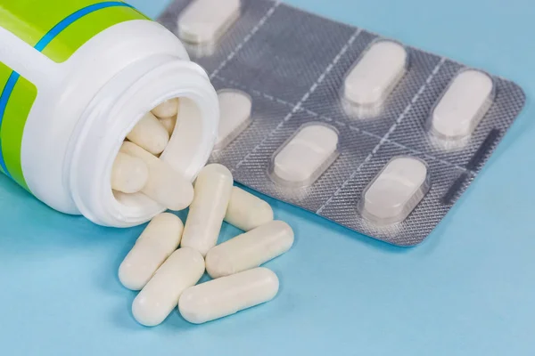 White capsules of the medication partly poured out from container in the form small plastic bottle on a blurred background of the blister pack of pills on the blue surface