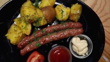 Kızarmış sosis, fırında patates ve siyah tabakta sos.