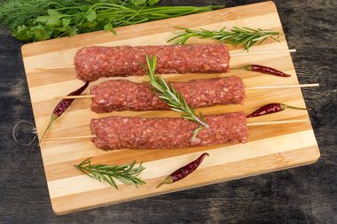 Tahta kesme tahtasının üzerinde, siyah bir yüzey üzerindeki sebzelerin, baharatların ve yeşilliklerin arasında birkaç çiğ lula kebabı şişlendi.