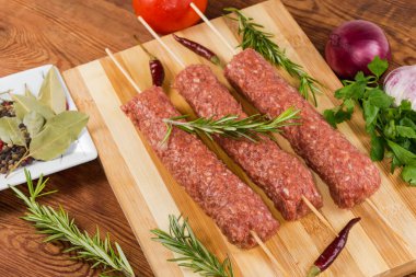 Birkaç çiğ lula kebabı, ahşap kesme tahtasındaki şişler üzerinde, sebzeler, baharatlar ve sebzeler arasında, eski kırsal masada, yakın plan.