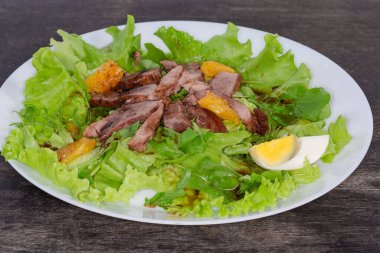 Kaynamış ördek eti, yumurta, portakal dilimleri ve siyah bir yüzeyde beyaz bir tabakta farklı yeşilliklerden yapılmış salata.