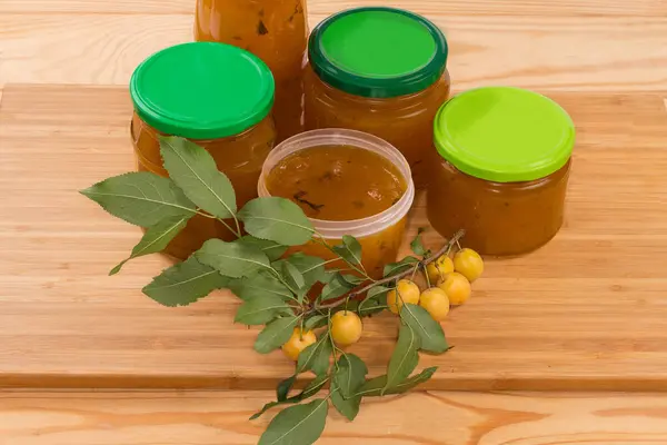 Yellow cherry plums jam made with mint leaves addition in open plastic container and several lidded jars, twig of the cherry plum tree with fruit on a cutting board