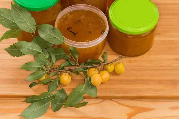 Yellow cherry plums jam made with mint leaves addition in open plastic container and several lidded jars, twig of the cherry plum tree with fruit on a cutting board, top view