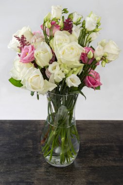 Beyaz ve pembe güller buketi ve cam şeffaf vazoda beyaz Eustoma, açık arkaplanda siyah bir yüzey üzerinde duran su.