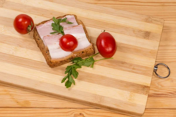 Open sandwich with slices of the boiled-smoked pork belly on skin and cherry tomatoes on the cutting board on the rustic table