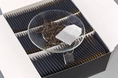 Kurumuş siyah çay yaprakları ve çay poşeti açık kağıt kutunun üzerinde, aynı çay poşetleriyle küçük kağıt paketlerde.
