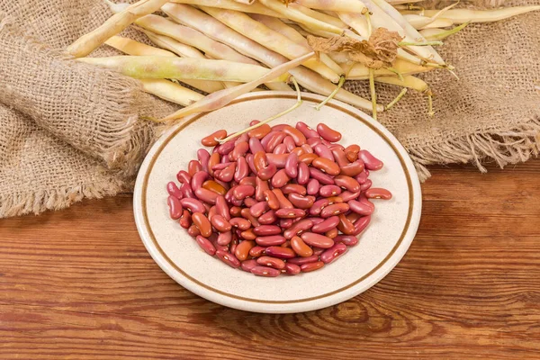 Raw husked red kidney beans on the saucer against the same beans in the pods on burlap on the rustic table