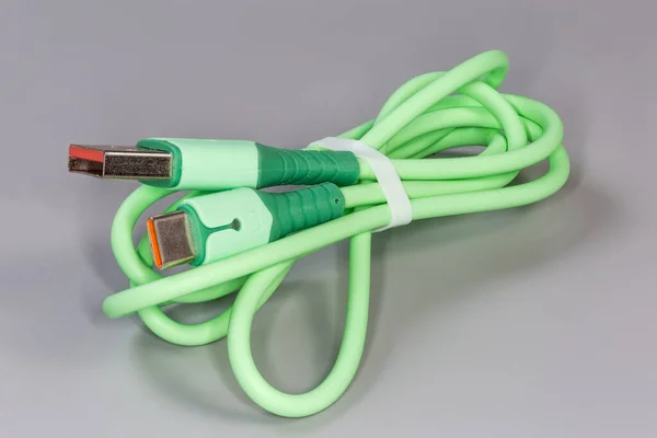 Twisted green USB cable with plugs standard A and standard C at the edges on a gray surface, close-up in selective focus