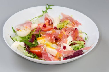 Jambon dilimlerinden yapılmış salata, haşlanmış bıldırcın yumurtası, taze yeşiller ve sebzeler beyaz bir tabakta, seçici odak noktasında gri bir arka planda yan görünüm