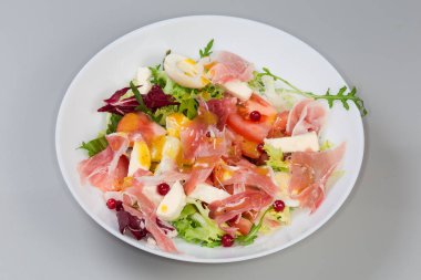 Jambon dilimlerinden yapılmış salata, haşlanmış bıldırcın yumurtası, taze yeşiller ve sebzeler gri arka planda beyaz bir tabakta.