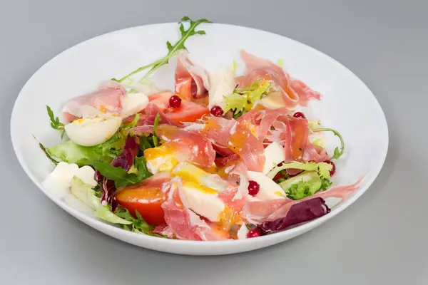 Jambon dilimlerinden yapılmış salata, haşlanmış bıldırcın yumurtası, taze yeşiller ve sebzeler beyaz bir tabakta, seçici odak noktasında gri bir arka planda yan görünüm