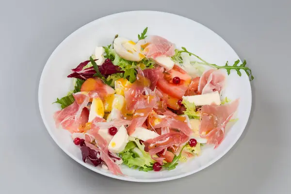 Jambon dilimlerinden yapılmış salata, haşlanmış bıldırcın yumurtası, taze yeşiller ve sebzeler gri arka planda beyaz bir tabakta.