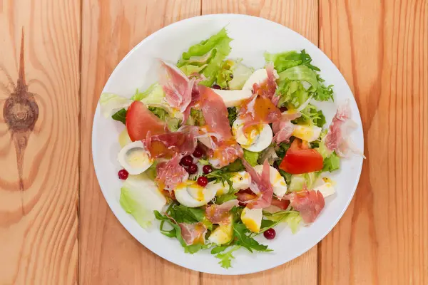 Jambon dilimlerinden yapılmış salata, kaynamış bıldırcın yumurtası, taze yeşiller ve sebzeler kırsal bir masada beyaz bir tabakta, üst tarafta.