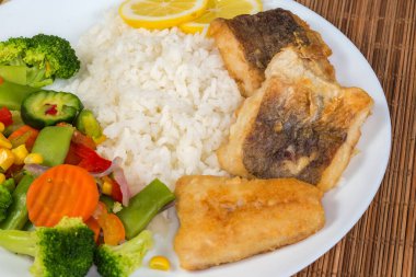 Kızarmış hake fileto parçaları haşlanmış sebzelerle garnitür karışımı ve pilav beyaz bir tabak üzerinde bambu yeri paspası, yakın plan.