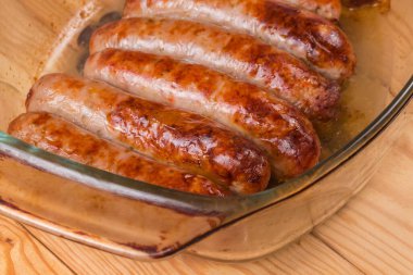 Baked thick pork sausages in natural casing on the old glass casserole pan on a rustic table, fragment close-up in selective focus