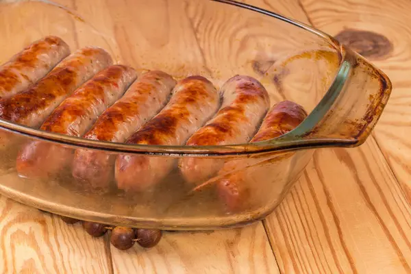 stock image Baked thick pork sausages in natural casing on the old glass casserole pan on a rustic table, fragment close-up in selective focus