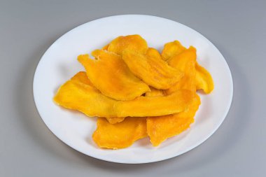 Gri arka planda beyaz tabakta kurutulmuş mango dilimleri.