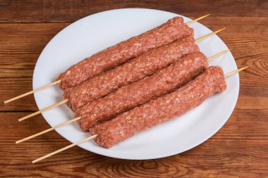 Birkaç çiğ soğumuş lula kebabı ahşap bambu şişlerinin üzerinde eski bir kırsal masada beyaz bir tabakta.