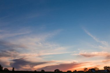 Gökyüzünün bir kısmı sirrus bulutlarıyla kaplı. Yazın gün batımında ufukta ağaç siluetleri ve tarım binaları var.