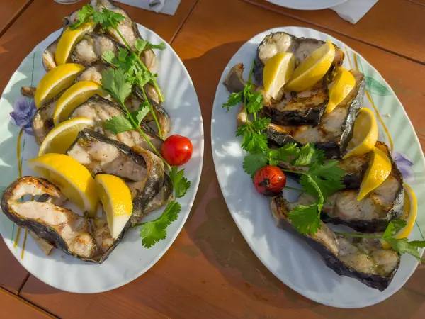 stock image Grilled steaks of the fresh Siberian sturgeon with lemon slices and cilantro twigs on the two plates on a table, top view