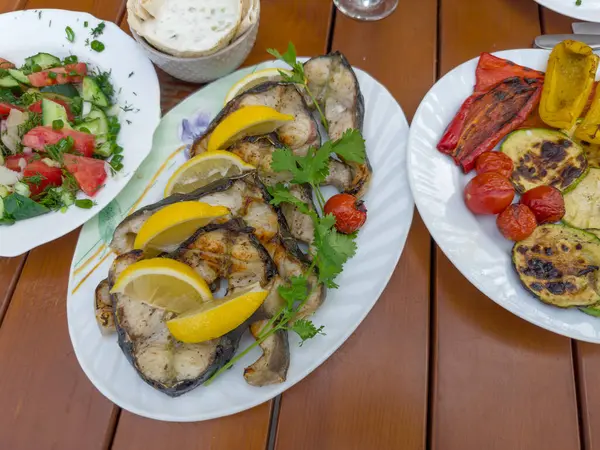 stock image Grilled steaks of the fresh sturgeon with lemon slices and cilantro twigs on the plate, salad of fresh vegetables, baked vegetables on a table, top view