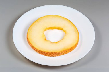 Olgun, taze, ağlı derili, sarı kavun parçası, gri arka planda beyaz bir tabakta kesiliyor.