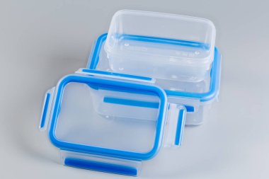 İki adet yeniden kullanılabilir şeffaf plastik gıda konteynırı farklı boyutlarda, biri gri arka planda mavi kapaklı.