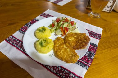 Tahta masadaki beyaz tabakta patates püresi ve taze sebze salatasıyla hamurda kızartılmış kemiksiz domuz pirzolası servis ediliyor.