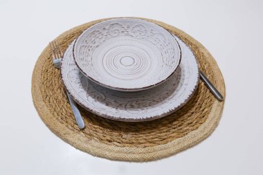 Derin kaseden gelen seramik beyaz-kahverengi sofra takımı ve yığının içinde geniş sığ tabak, beyaz bir masanın üzerindeki paslanmaz çelik çatal bıçak seti.