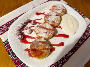 Serving of the several fried cottage cheese pancakes watered with raspberry jam and sprinkled with sugar powder, sour cream and fresh raspberry berries on dish on the wooden table, top view clipart