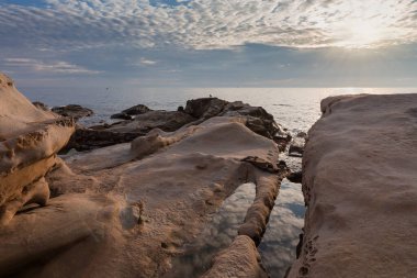 Bulutlu gökyüzünün arka planında taştan kıyı şeridi olan deniz kıyısının bir kısmı ve güneşli bir sabahta bulutların arkasından gelen güneş ışınları.
