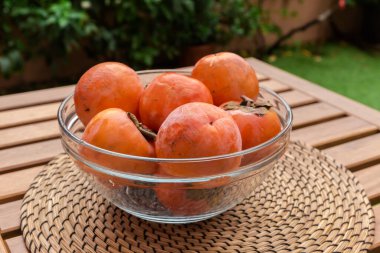 Şeffaf cam mutfak kâsesinde, ahşap masada açık havada servis altlığı olan olgun hurma meyveleri.