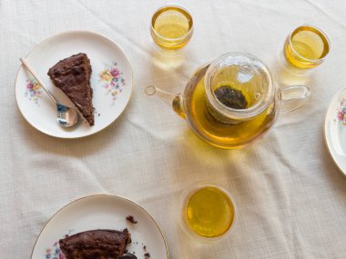 Üç şeffaf cam fincanda bitkisel karışım ile taze demlenmiş yeşil çay ve masa örtüsüyle kaplı bir masada çay tabağının üzerinde kek parçaları olan cam çaydanlık.