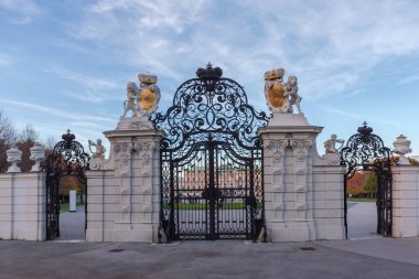 Central forged gate with two open side wickets in the Upper Belvedere against the partly cloudy sky and palace at sunset in autumn, Vienna, Austria, view from outside clipart