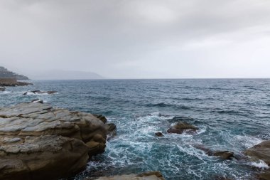 Bulutlu gökyüzünün arka planında taştan kıyı şeridi olan deniz kıyısının bir kısmı ve bulutlu bir günde sisli kıyı tepeleri.